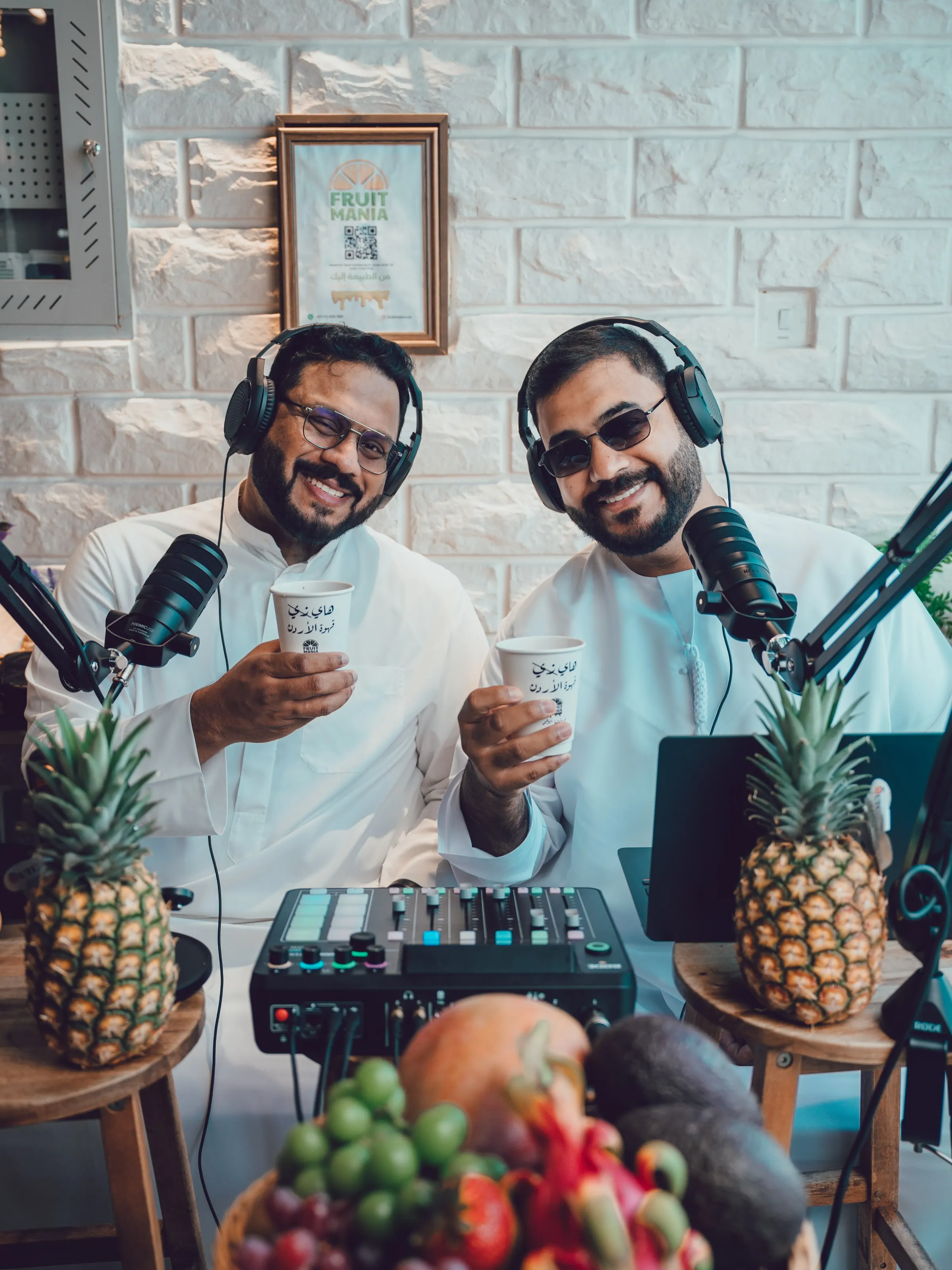 Two presenters in white kanduras wearing headphones at podcast microphones, with an audio mixer and a bowl of fruit on the table